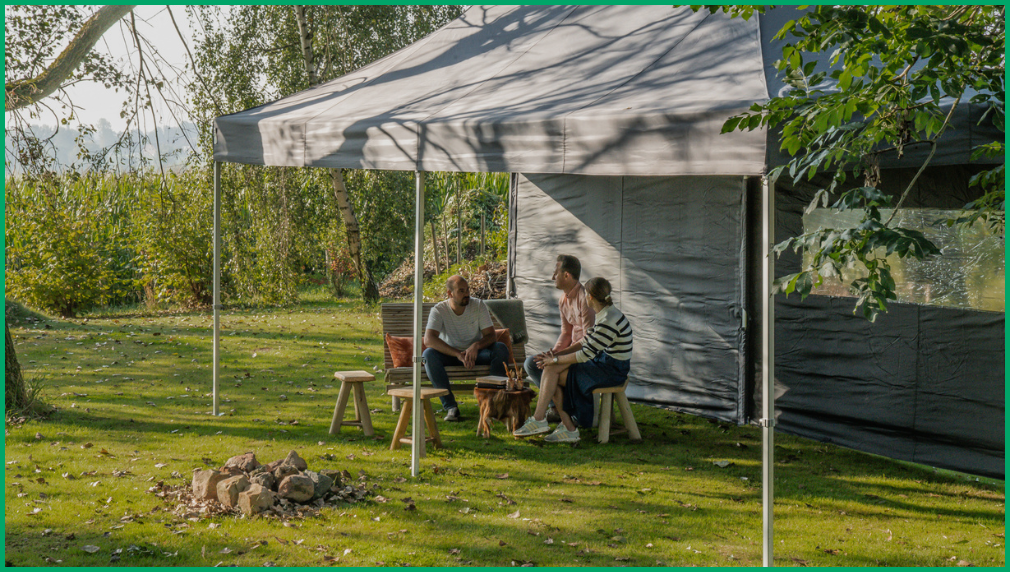 Familie zittend aan picknicktafel onder een witte vouwtent in een groene tuin omgeven door bomen