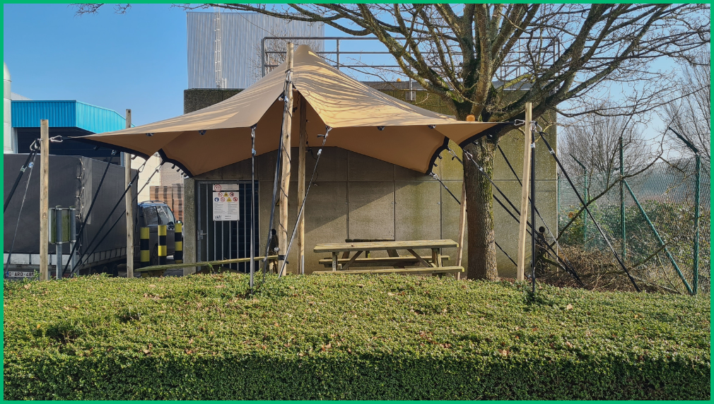 Beige stretchtent op houten palen bij industrieel bedrijfsgebouw met picknicktafel eronder