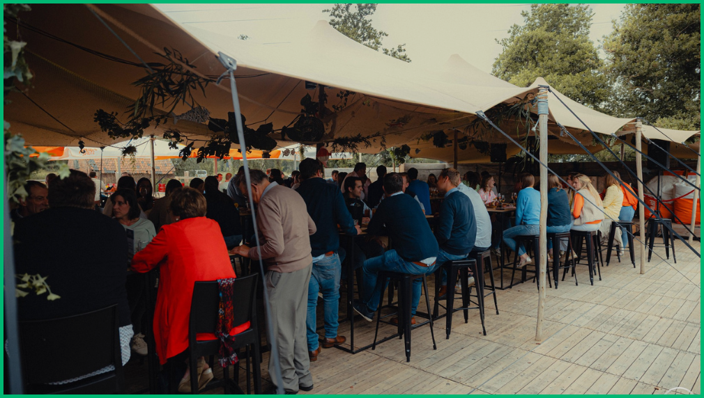 Mensen zittend aan hoge tafels onder een witte stretchtent bij SDE beurs netwerkevenement buiten